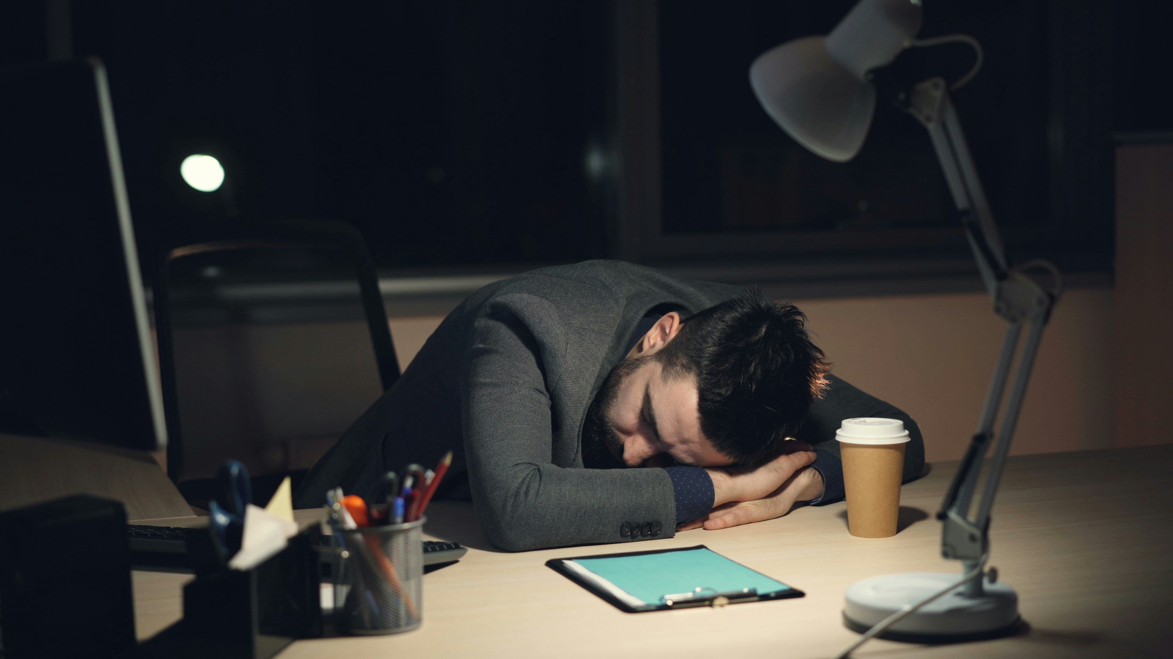 Person sleeping at a desk with a laptop, lamp, and coffee cup.